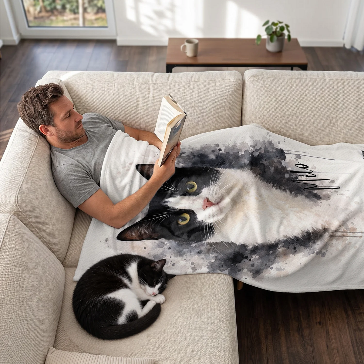 Mann liest auf dem Sofa unter der Aquarell-Katzendecke, daneben schläft seine Katze
