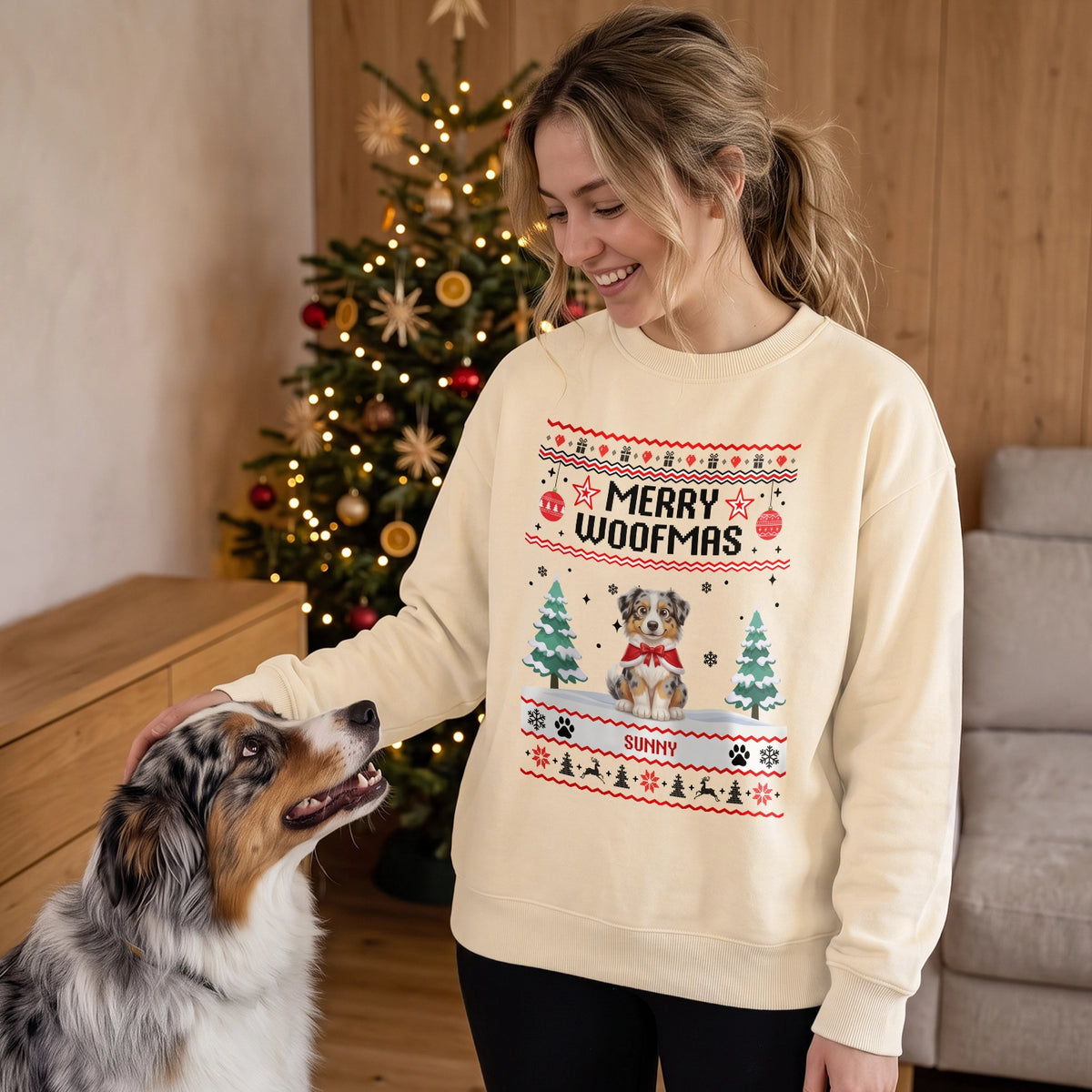 Frau im hellbeigen Merry-Woofmas-Pullover mit ihrem Australian Shepherd vor dem Weihnachtsbaum